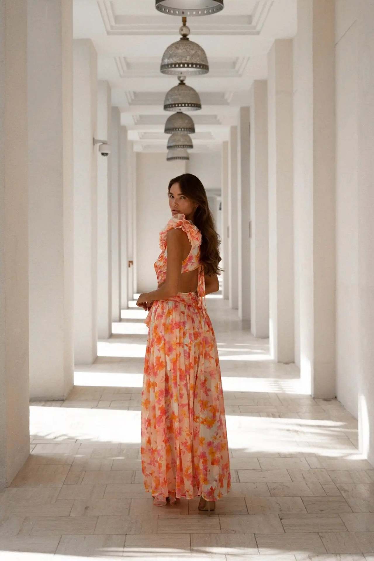 Woman in an orange floral dress standing in a sunlit hallway with columns.