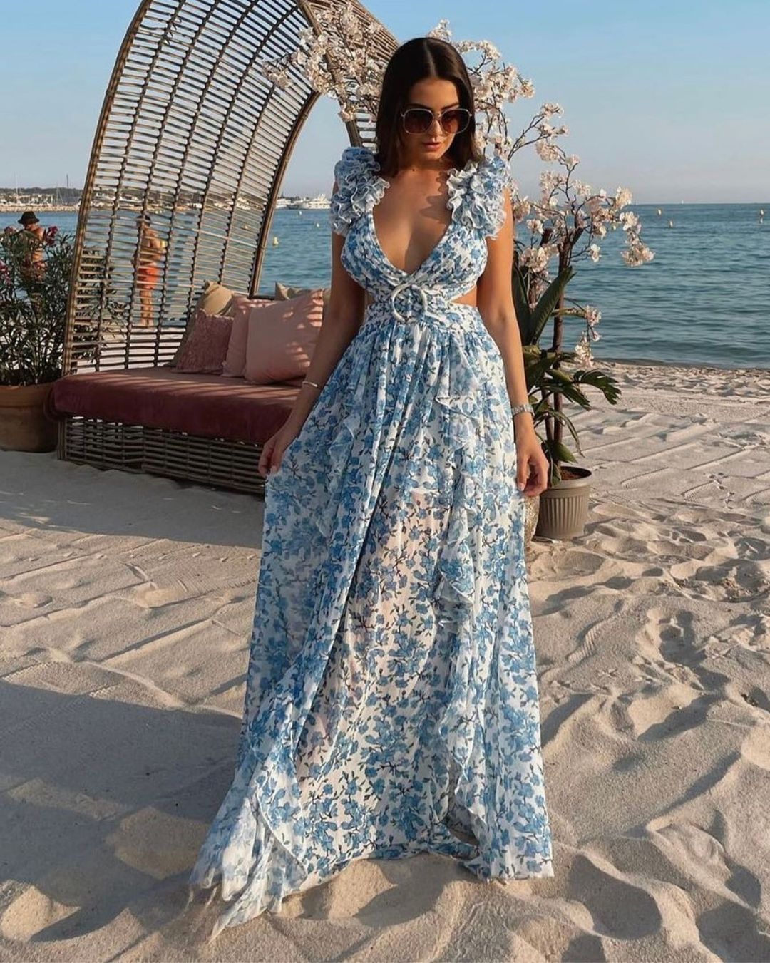Woman in a blue floral dress standing on a sandy beach with ocean view.