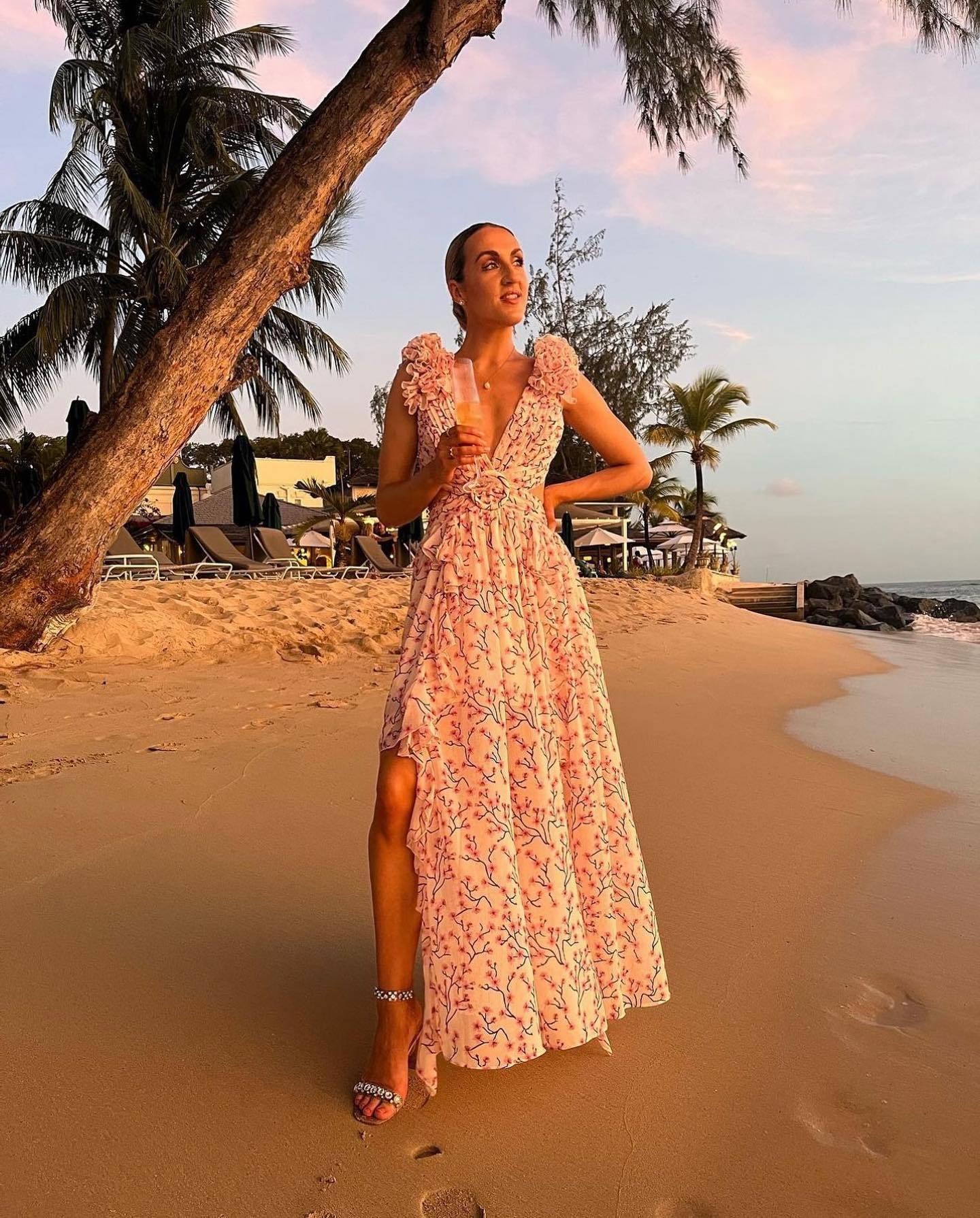 Woman in a floral dress standing on a beach with palm trees and ocean view.