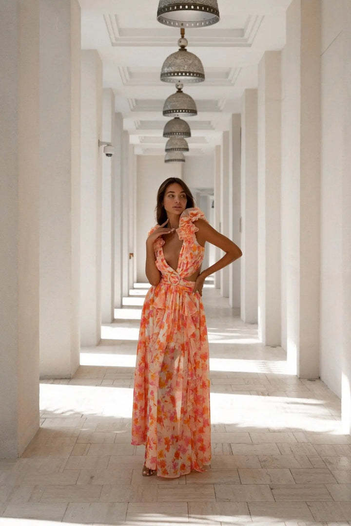 Woman in an orange floral dress standing in a sunlit hallway with columns.