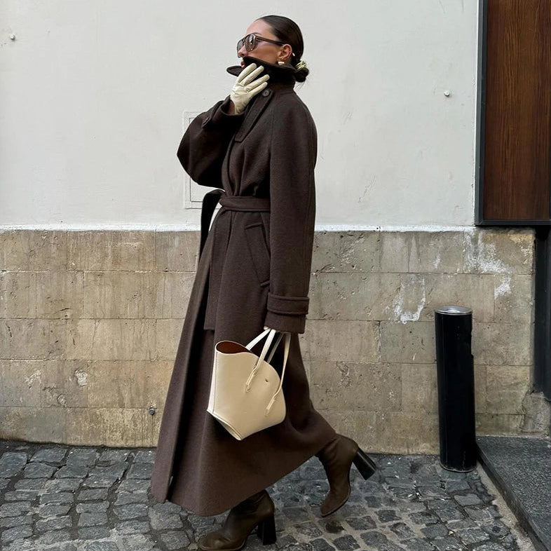Woman in a long brown coat walking on a street, holding a beige handbag.