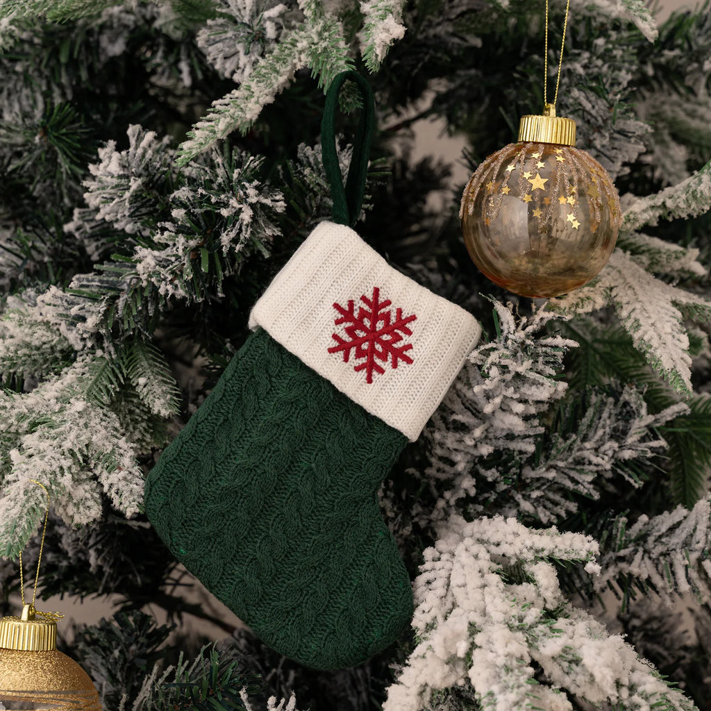 Green knitted Christmas stocking with a red snowflake design hanging on a decorated tree.
