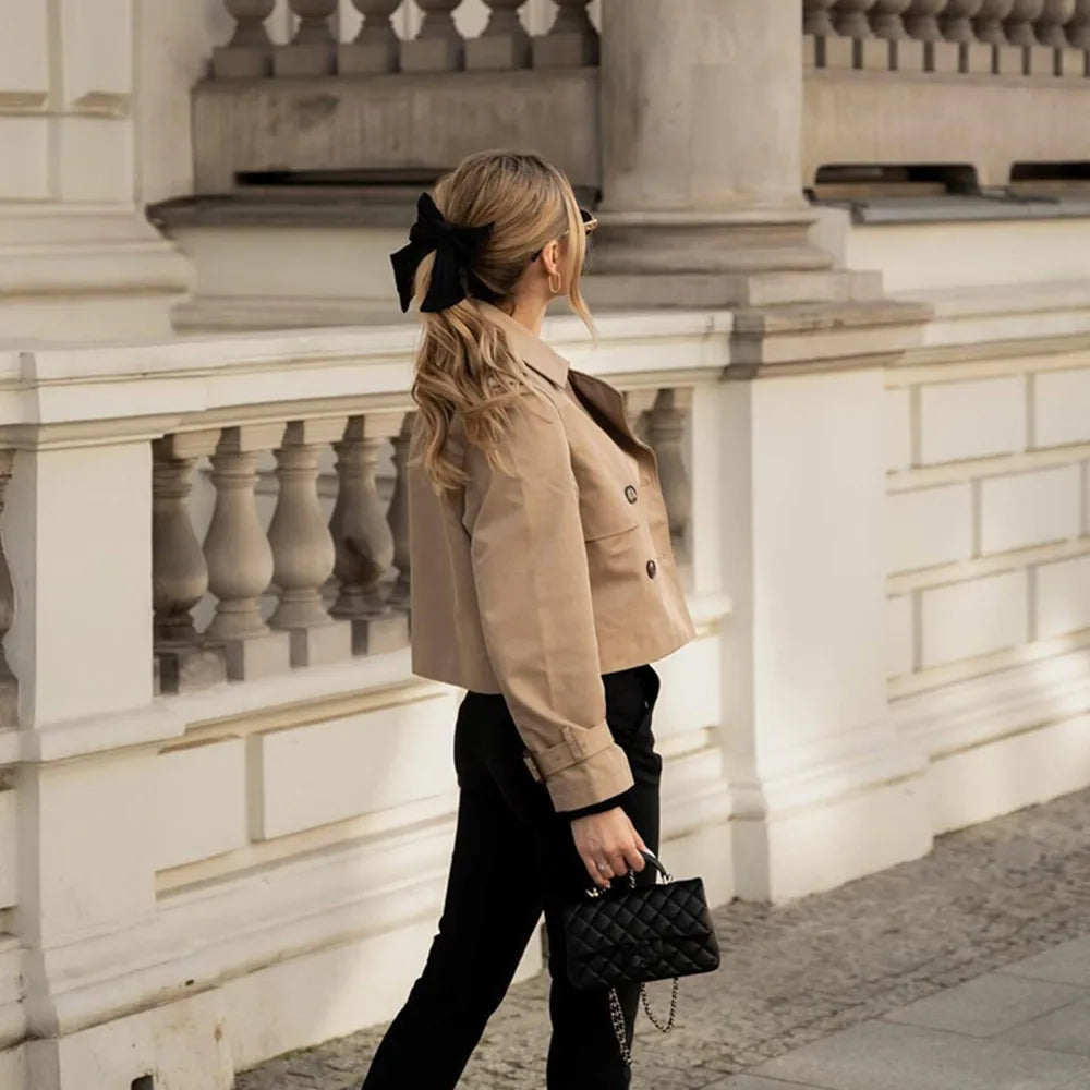 Woman in a beige jacket and black pants walking away from the camera with a decorative hair bow.
