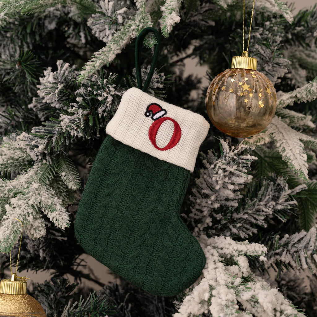 Green knitted Christmas stocking with a red letter 'O' and white cuff hanging on a decorated tree.