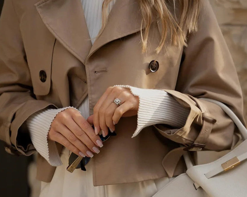 Person wearing a beige trench coat holding a white handbag against a stone wall background