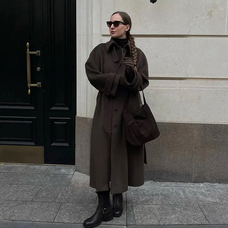 Woman in a long brown coat standing on a sidewalk next to a building entrance.