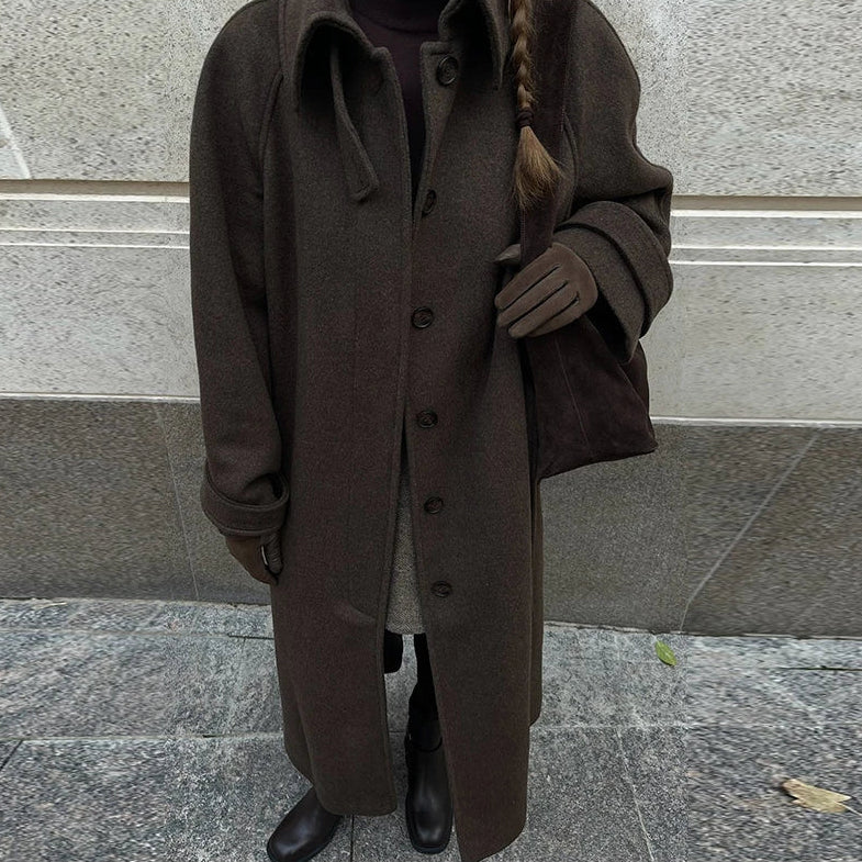 Person wearing a long brown coat standing against a stone wall.