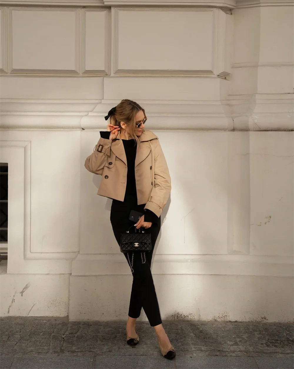 Woman in beige coat and black outfit standing against a white wall.