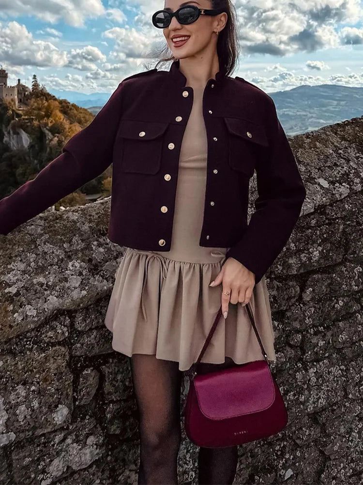 Woman in a dark jacket and beige skirt holding a purple bag against a scenic background.