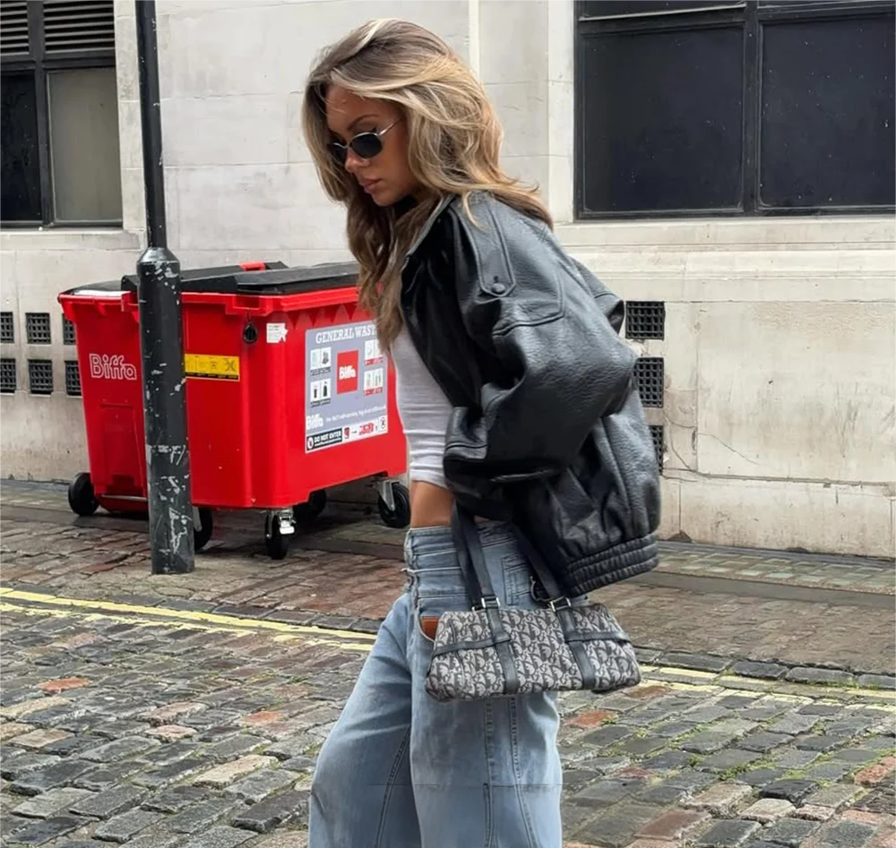 Woman in a leather jacket and sunglasses walking on a street with a red bin in the background.