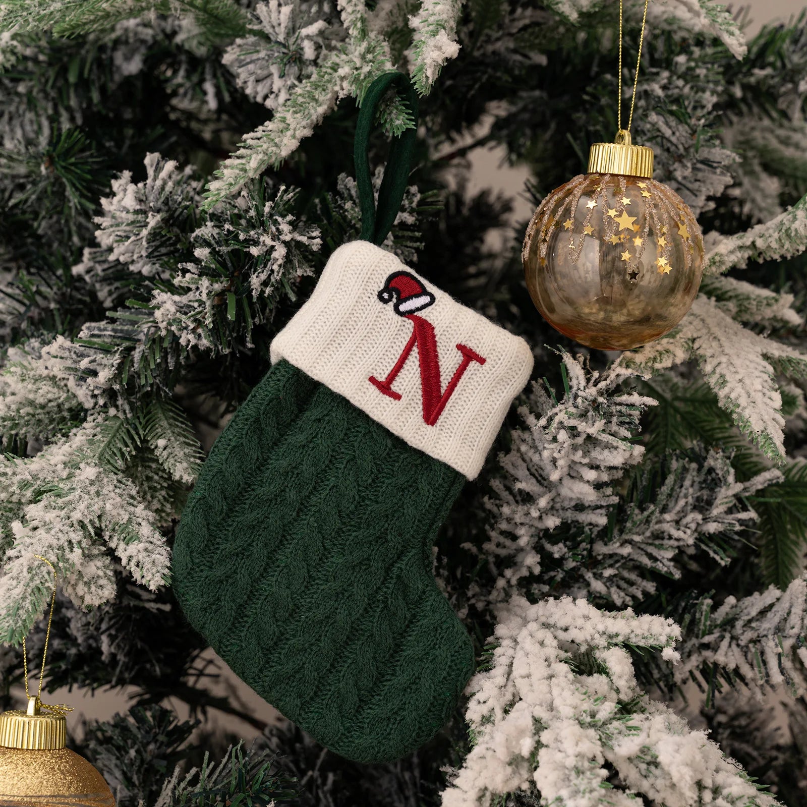 Green knitted stocking with a red 'N' ornament hanging on a Christmas tree.