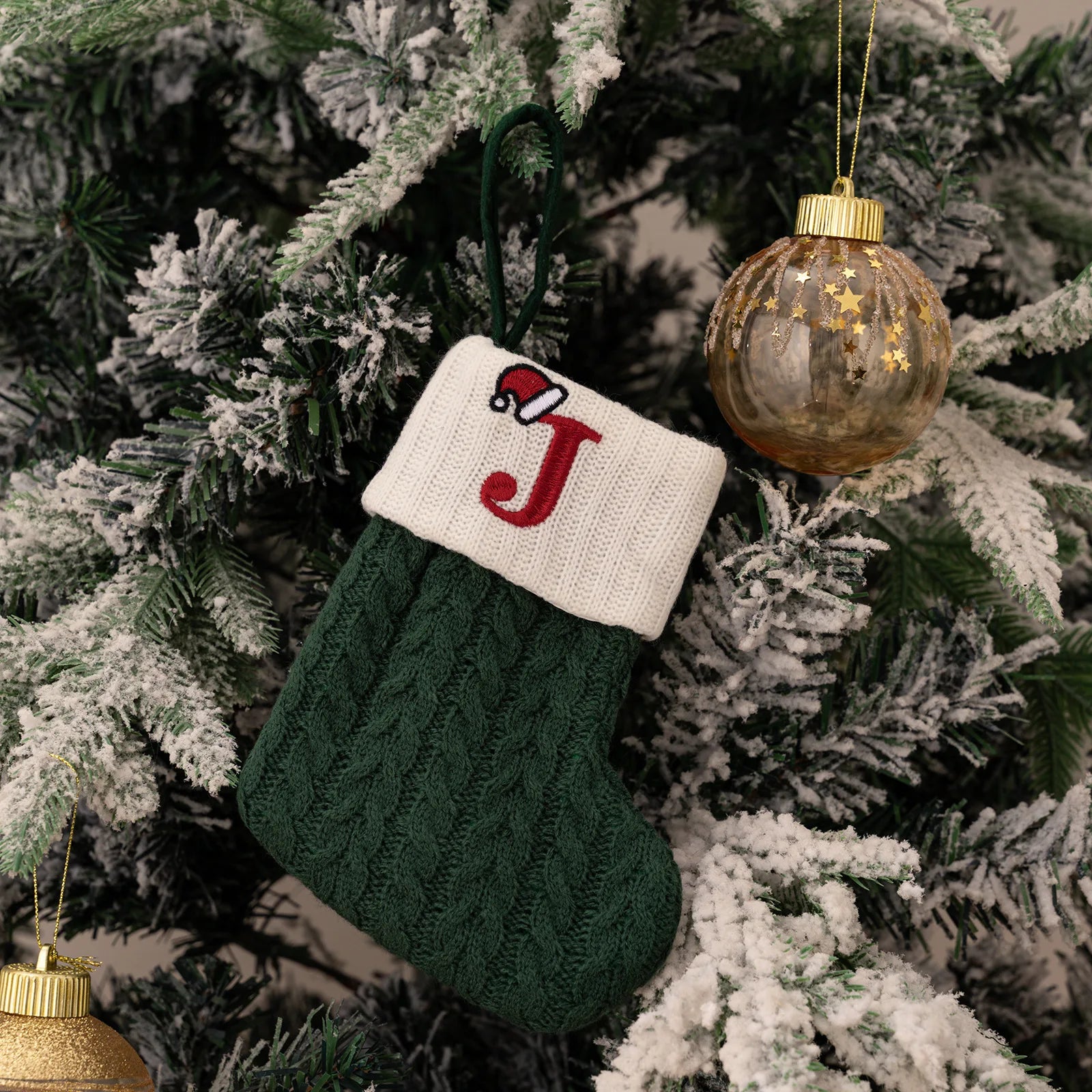 Green knitted stocking with a red letter 'J' ornament hanging on a decorated Christmas tree.