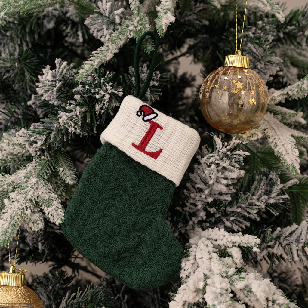 Green knitted stocking with a red 'L' hanging on a decorated Christmas tree.