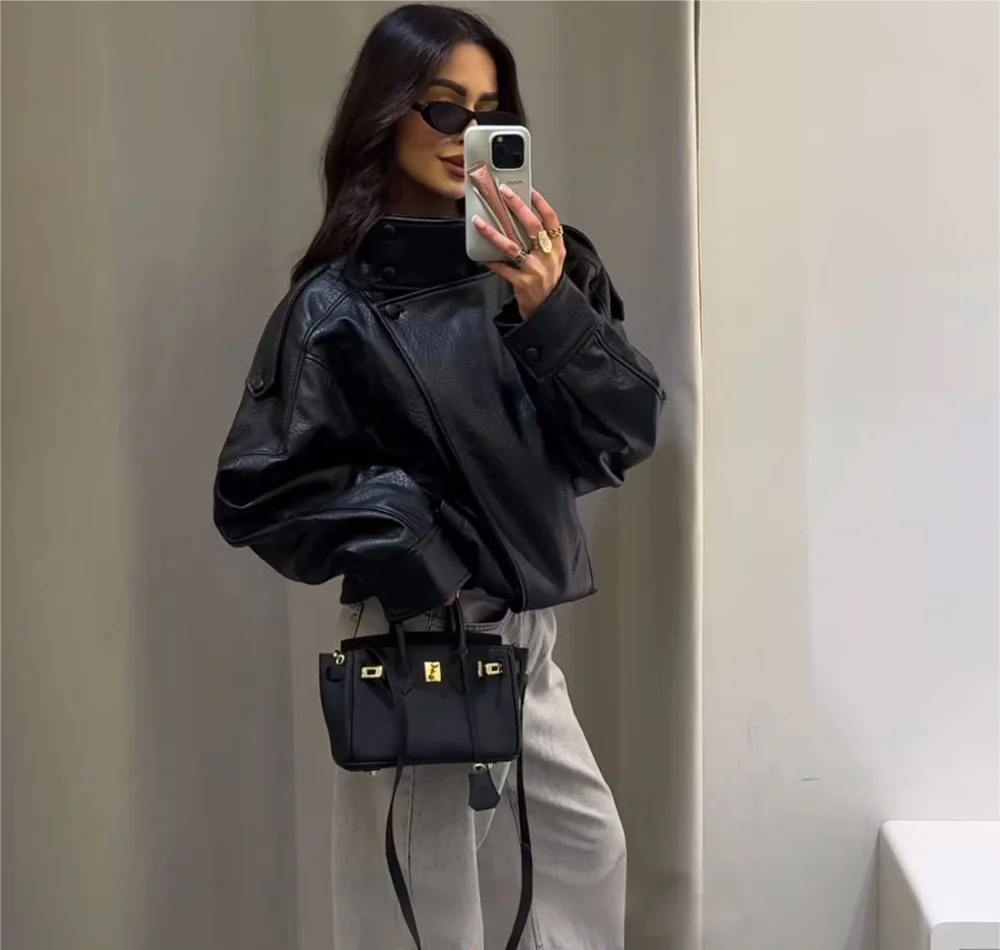 Person wearing a black leather jacket and holding a black handbag, taking a mirror selfie.