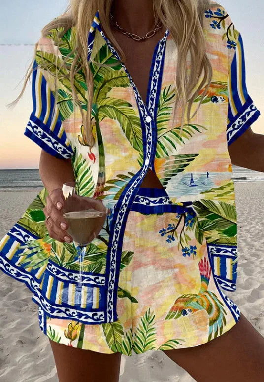 Woman in a tropical print outfit holding a drink on a beach.