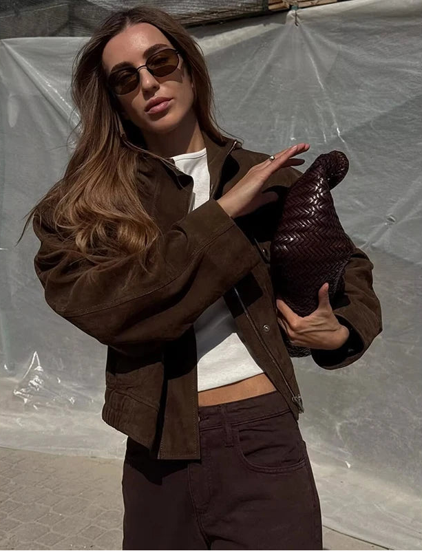 Woman wearing a brown jacket and sunglasses, holding a brown bag against a plain background