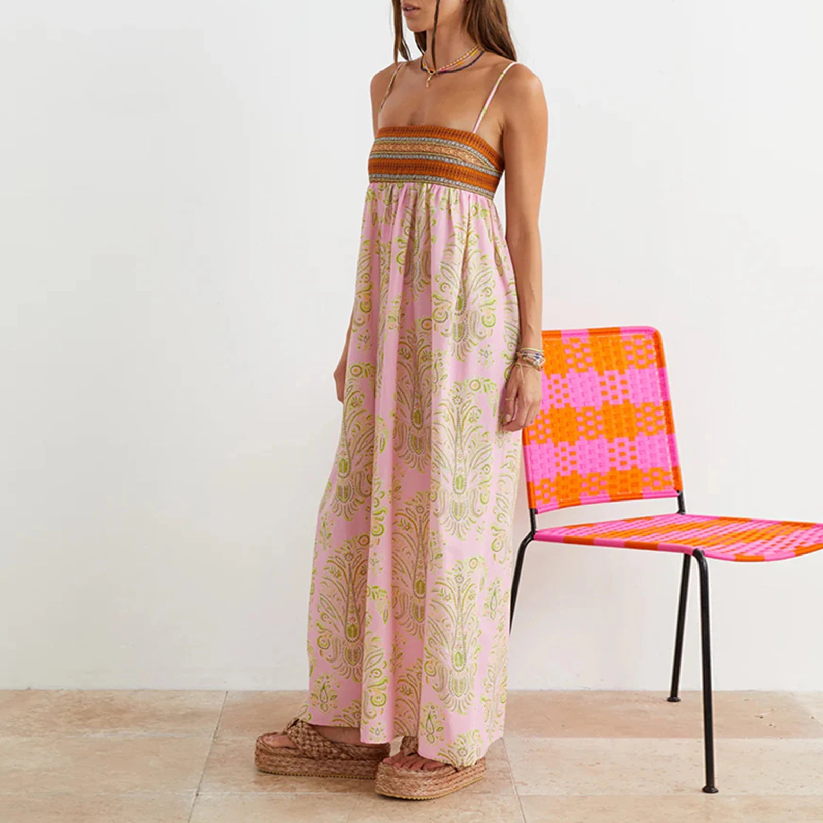 Woman in a long pink dress standing next to a colorful chair on a wooden floor.