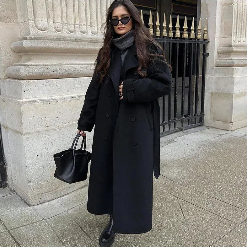 Woman in a long black coat standing in front of a stone building.