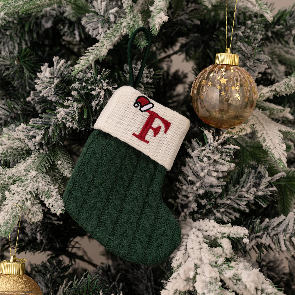 Green knitted Christmas stocking with a red letter 'F' on a decorated tree.