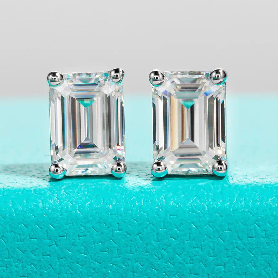 Pair of emerald-cut diamond earrings on a turquoise surface with 'Anu Jewel' branding.