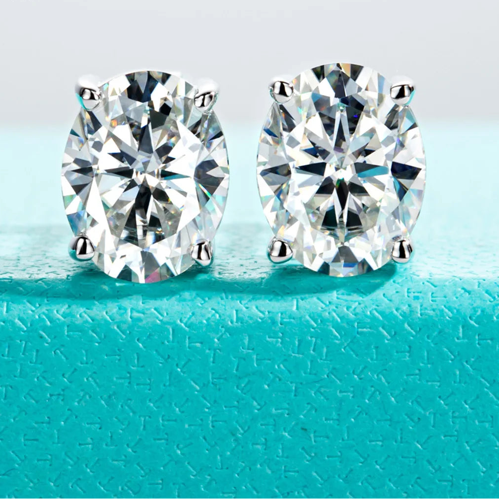 Pair of diamond stud earrings on a turquoise surface with 'Anu Jewel' branding.