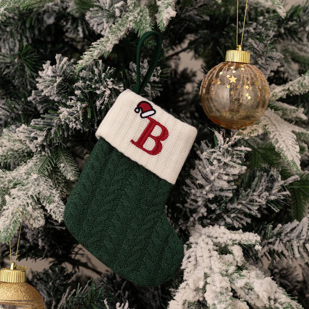 Green Christmas stocking with a red letter 'B' and white cuff, hanging on a decorated tree.