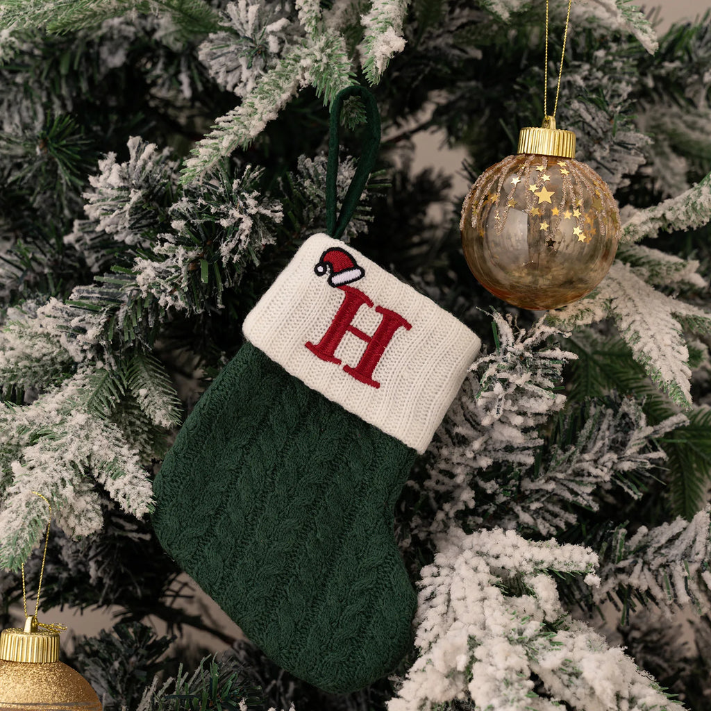 Green knitted stocking with a red 'H' ornament on a decorated Christmas tree.