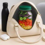 Beige handbag with a clear section revealing a jar of salad, next to a black water bottle and white device on a light surface.