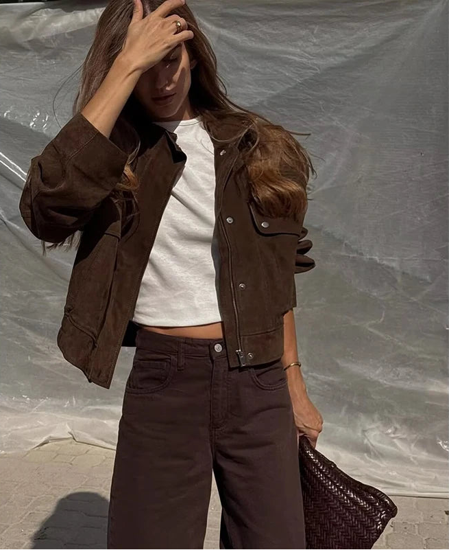 Woman wearing a brown jacket and pants with a white top, holding a woven bag against a plain background.