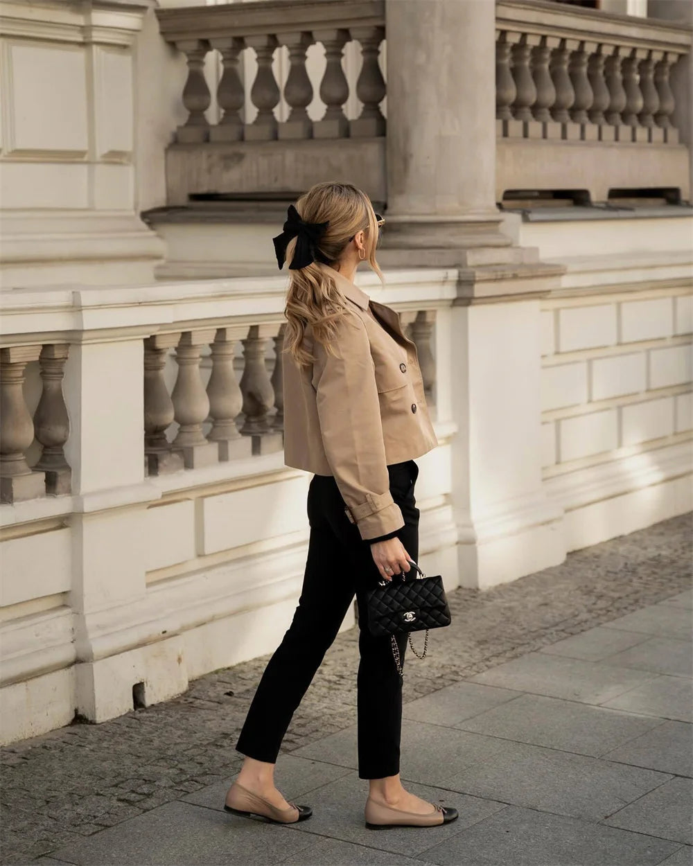 Woman in beige jacket and black pants walking on a sidewalk with a building in the background
