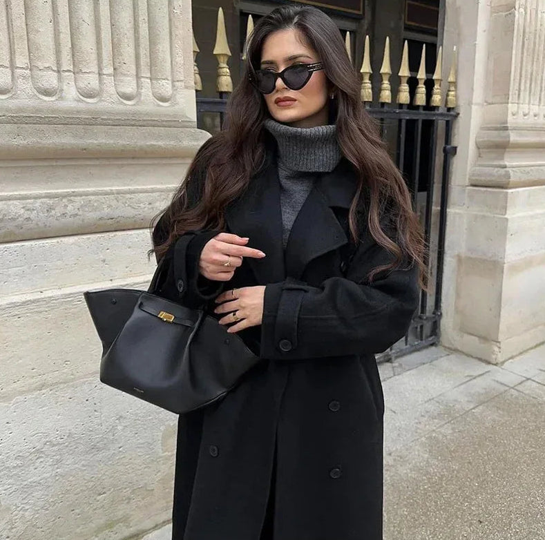 Woman in a black coat holding a black handbag in front of a stone building.