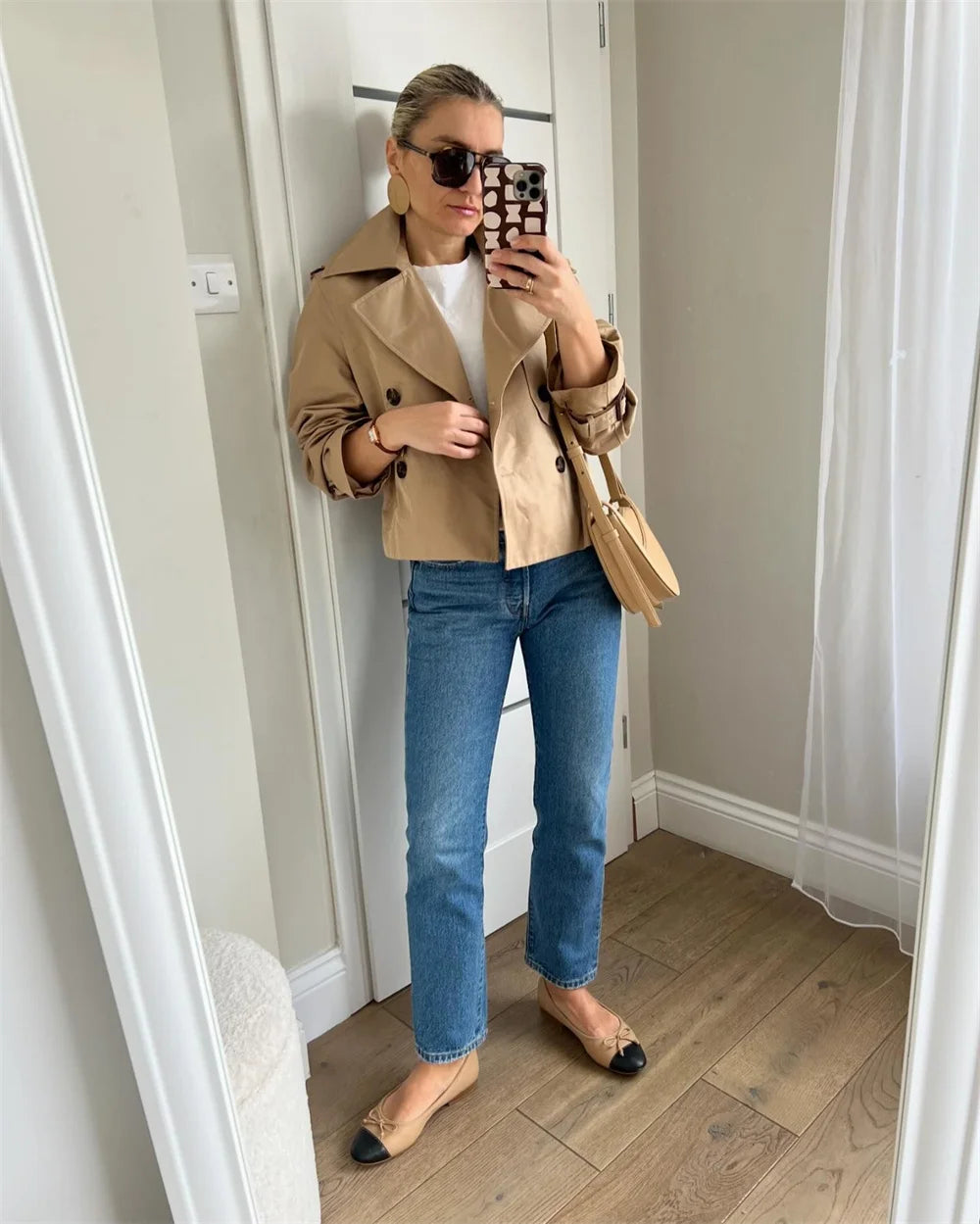 Woman taking a mirror selfie wearing a beige jacket, white shirt, blue jeans, and black shoes.
