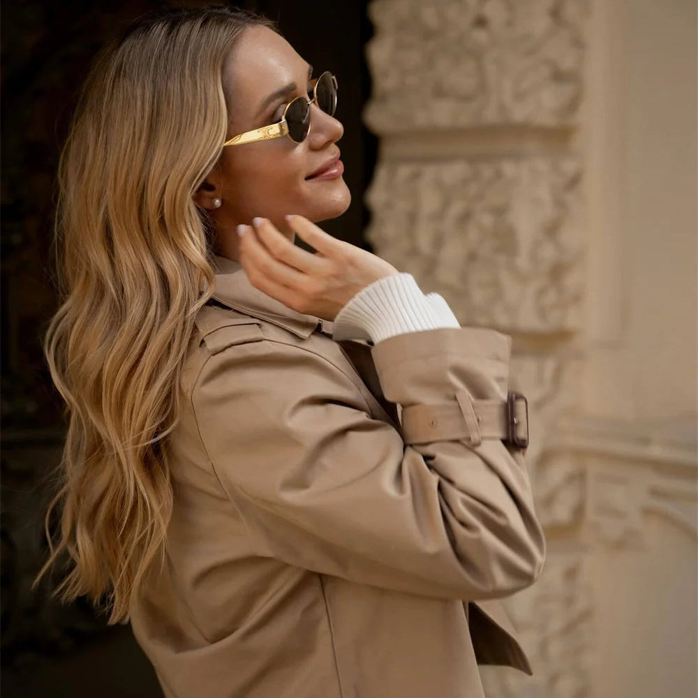 Woman wearing sunglasses and a beige coat in front of an architectural background