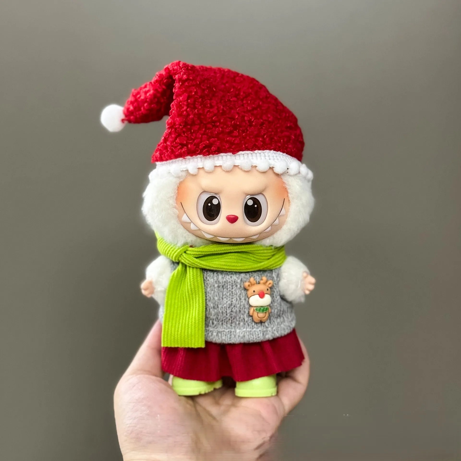 Small plush toy with a red Santa hat, green scarf, and gray sweater held in a hand against a plain background.