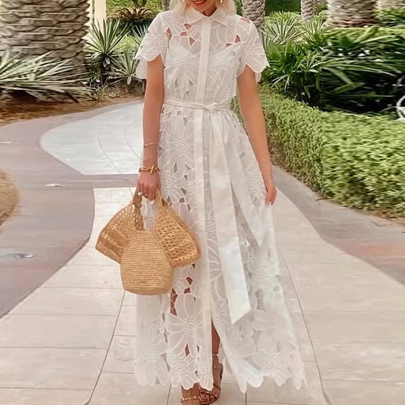 Woman in a white lace dress holding a straw bag on a pathway with greenery.
