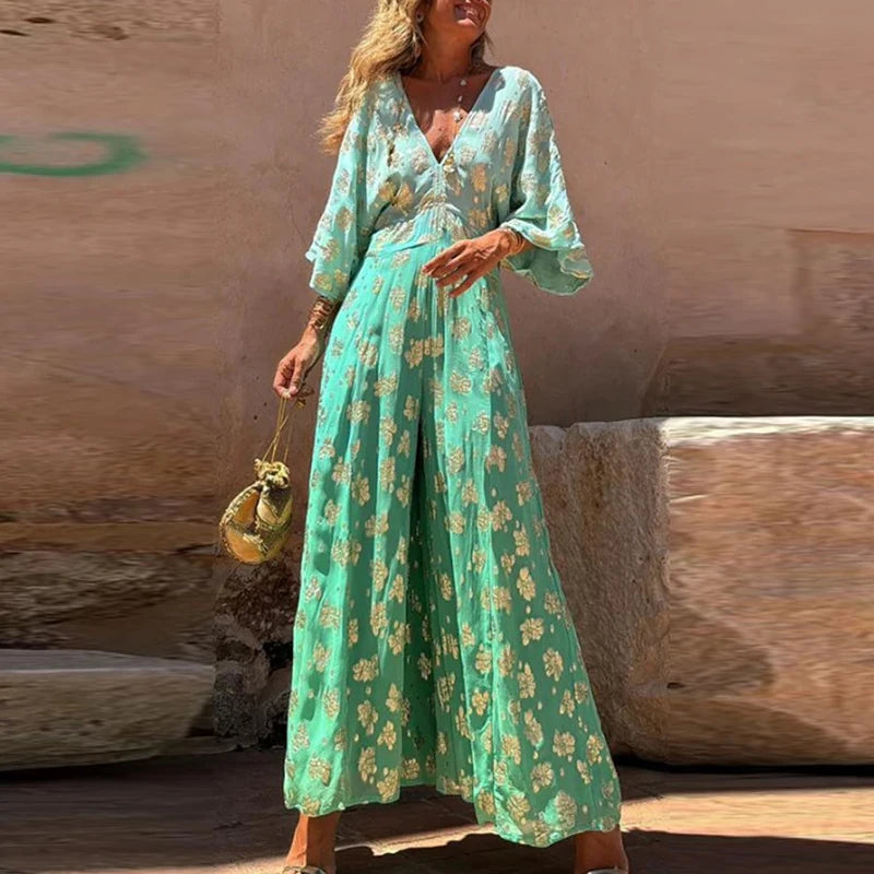 Woman in a long turquoise floral dress standing against a stone wall.