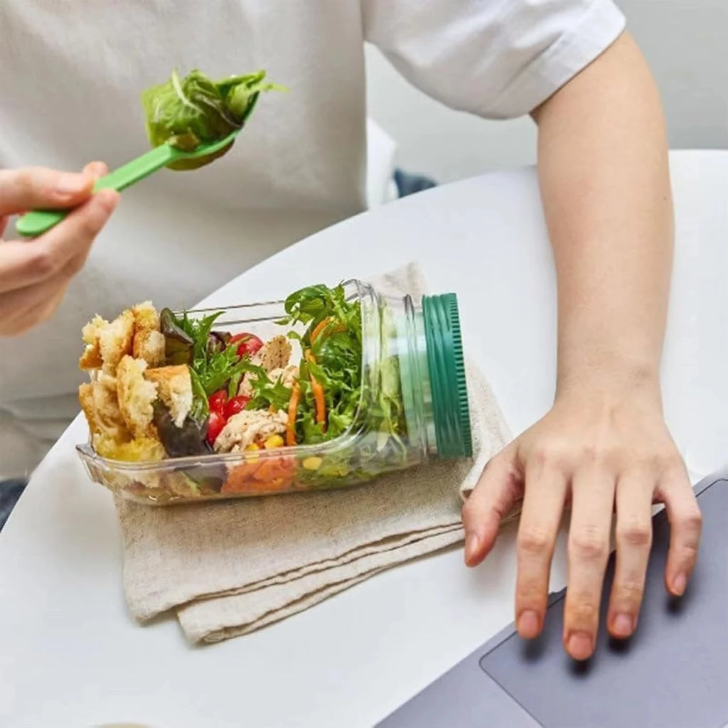Person holding a jar of salad with a side of fried food on a white surface.