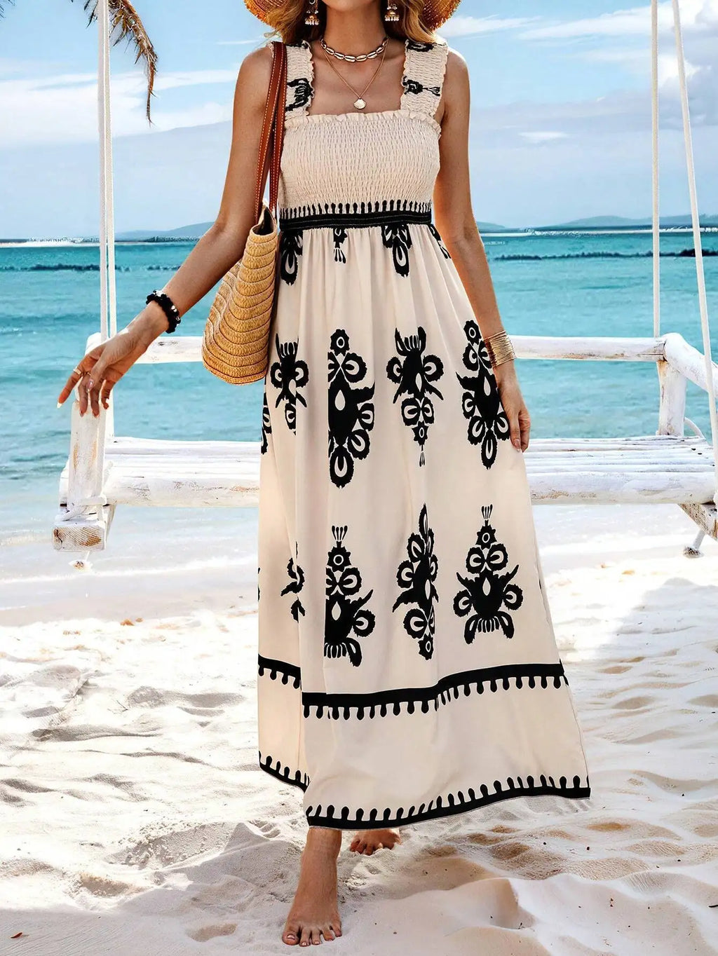 Woman in a long dress with black patterns standing on a sandy beach with ocean view.