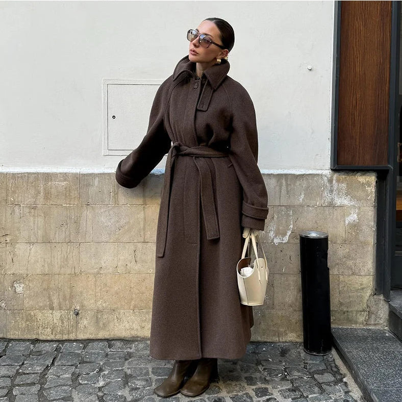 Woman in a long brown coat standing on a street corner.