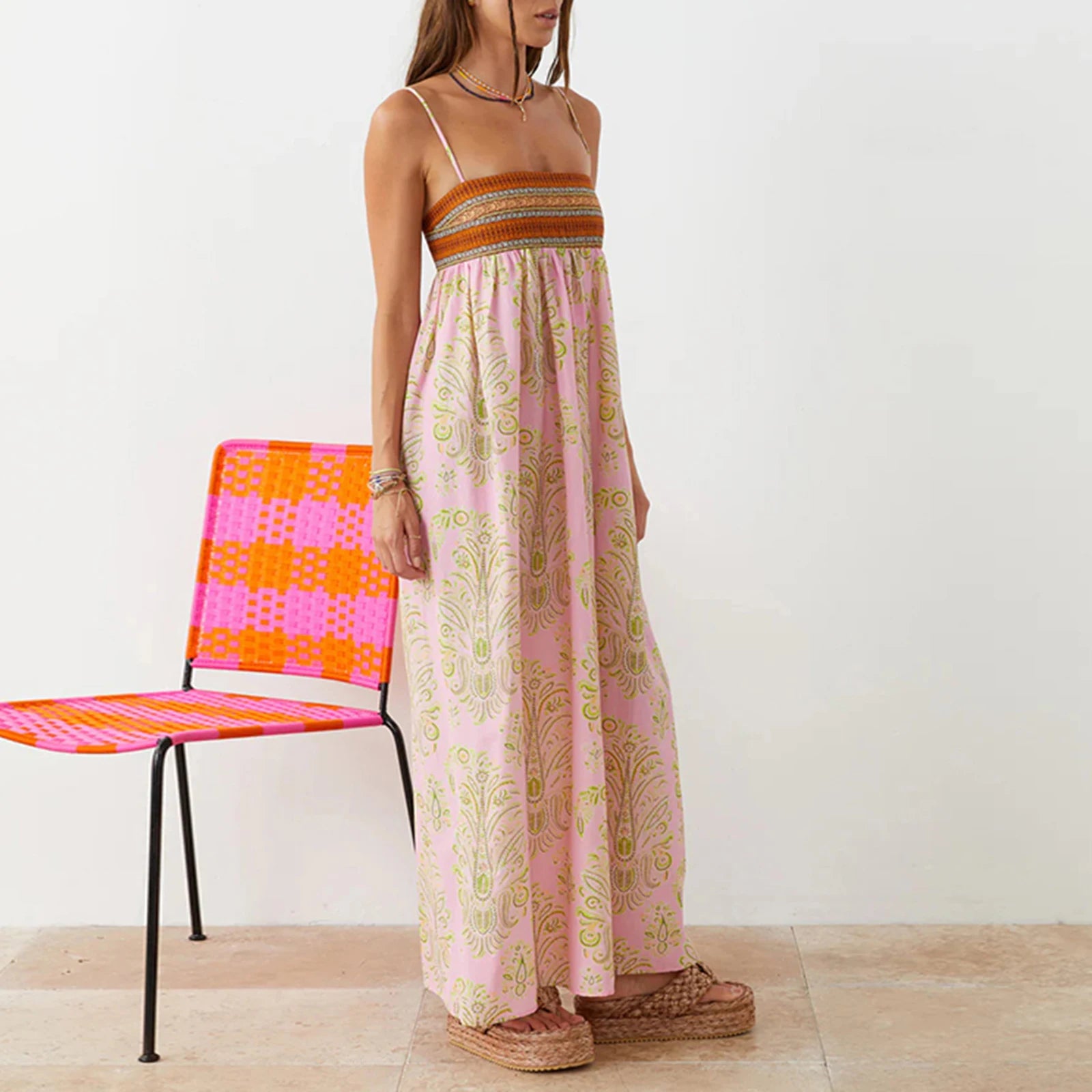 Woman wearing a long pink dress with a pattern, standing next to a colorful chair on a light wooden floor.