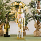 Decorative nutcracker figure in gold and white standing next to a bottle of wine with Christmas decorations in the background.