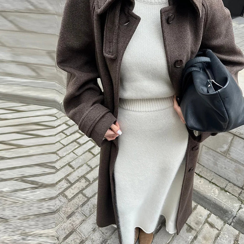 Person wearing a brown coat and white sweater holding a black bag on a stone pavement.