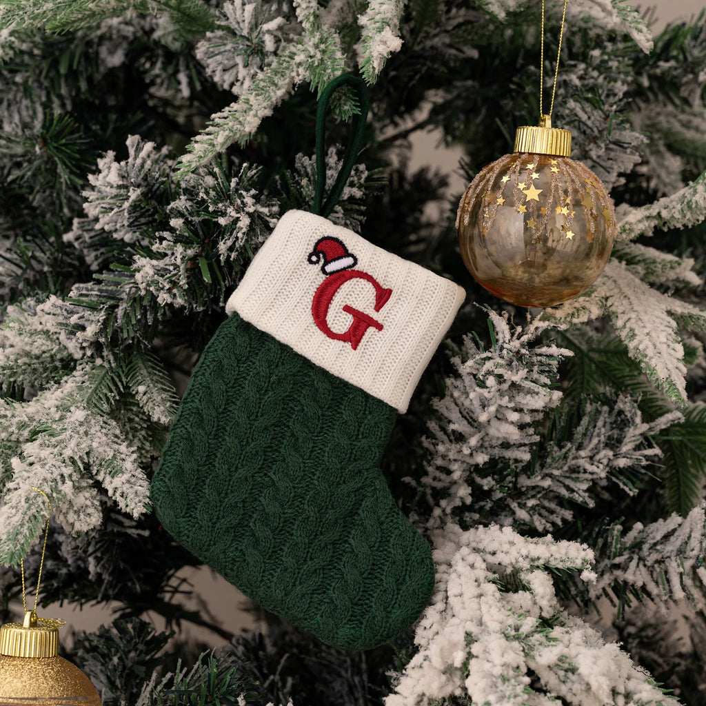 Green Christmas stocking with a white cuff featuring a red letter 'G' hanging on a decorated tree.