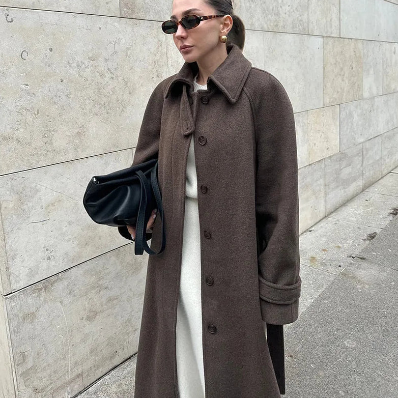 Woman wearing a brown coat and sunglasses, standing against a stone wall.