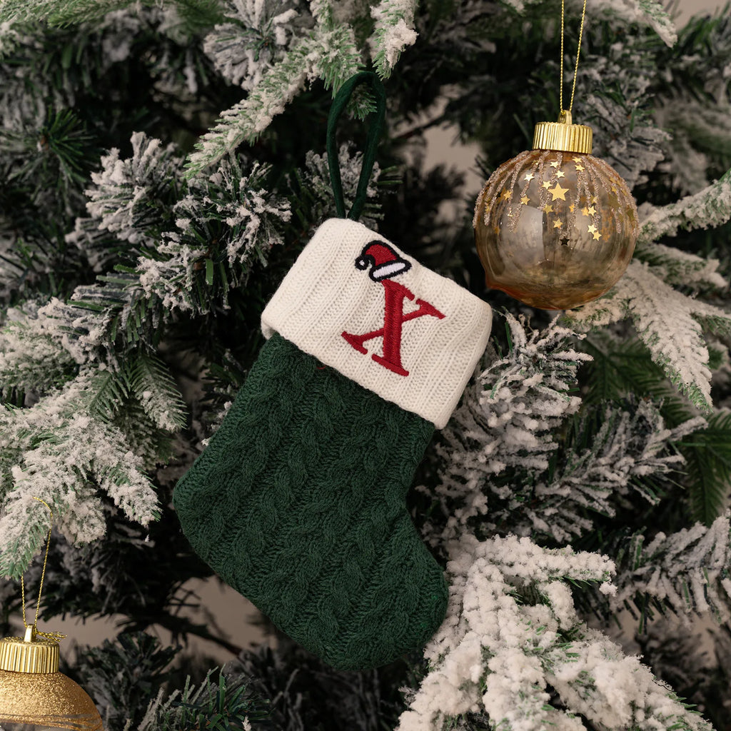 Green and white Christmas stocking with letter 'X' hanging on a decorated tree.